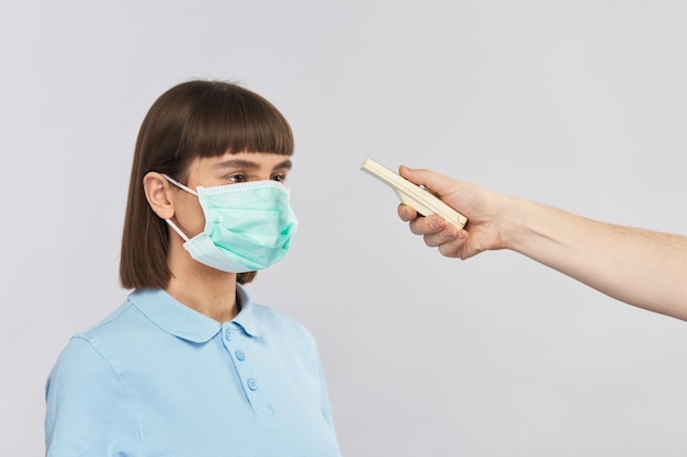 Mujer joven en máscara de protección de pie y brazo médico medir la temperatura con dispositivo electrónico