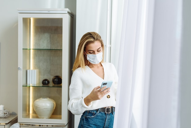 Una mujer joven con una máscara protectora está junto a la ventana y sostiene un teléfono en sus manos. Concepto de autoaislamiento, protección contra coronavirus.