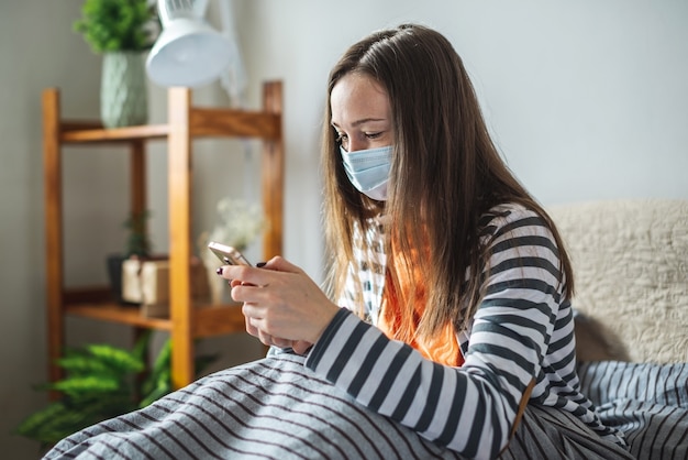 Mujer joven con una máscara protectora está sentada en su casa en la cama y usando su teléfono