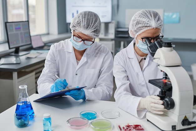 Mujer joven con máscara protectora y guantes examinando muestras con microscopio mientras su colega toma notas en la mesa