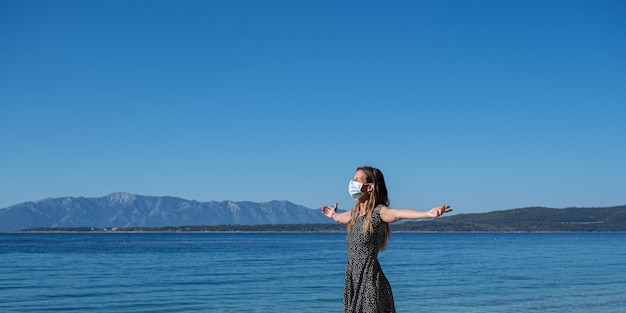 Mujer joven con máscara protectora médica en la cara de pie con los brazos extendidos por el mar en una imagen conceptual de la pandemia del coronavirus