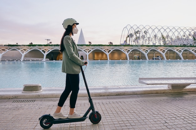 Mujer joven con máscara protectora montando un scooter eléctrico en la ciudad