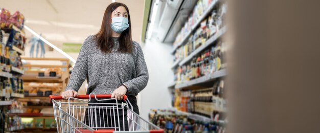 Una mujer joven con una máscara protectora en un supermercado camina con un carrito de compras y selecciona productos en los estantes Supermercado y compradores Lugar para su texto