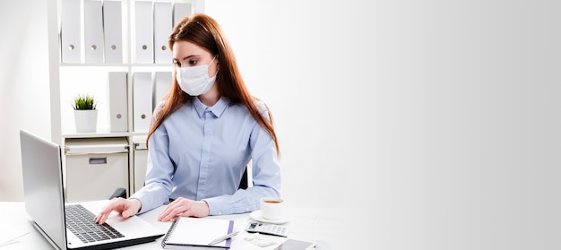 Una mujer joven con una máscara protectora trabaja en una computadora. Mujer de negocios en una máscara médica en la oficina.