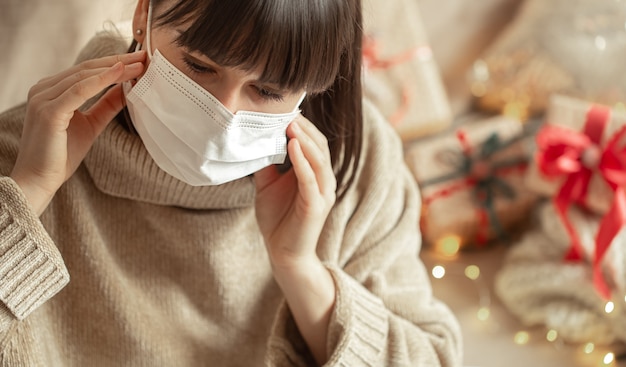 Mujer joven con una máscara en el rostro con un acogedor suéter beige en una pared borrosa con bokeh. El concepto de vacaciones de invierno durante el coronavirus.