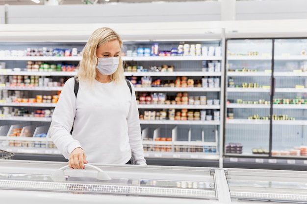 Mujer joven con una máscara en un supermercado elige alimentos congelados