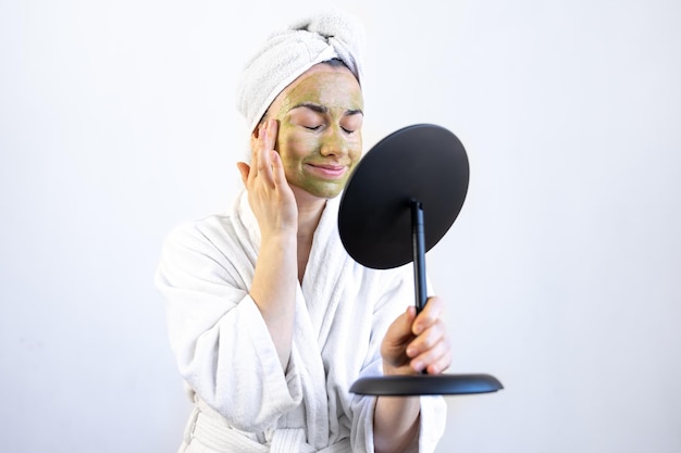 Mujer joven con una máscara verde en la cara en una bata de baño frente a un espejo