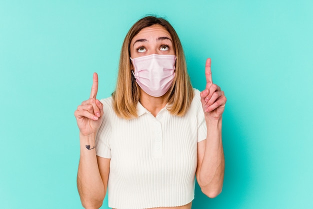 Mujer joven con una máscara para el virus aislado en la pared azul apuntando hacia arriba con la boca abierta.