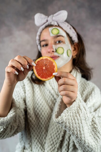 Mujer joven con mascarilla de arcilla facial y pepinos de pomelo