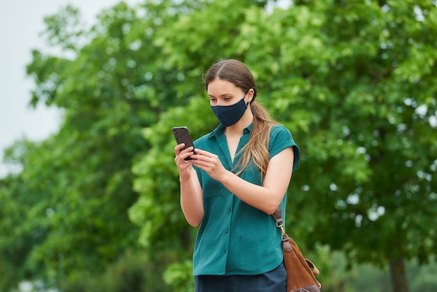 Una mujer joven con una mascarilla médica azul marino lee noticias en un teléfono inteligente mientras camina en el parque