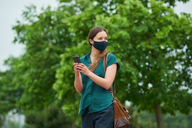 Una mujer joven con una mascarilla médica azul marino sosteniendo un teléfono inteligente con ambas manos mientras camina en el parque