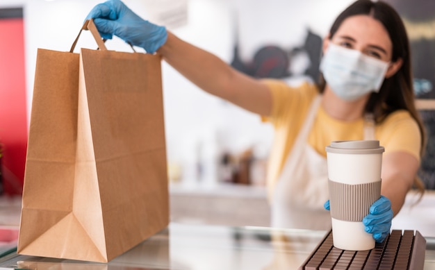 Mujer joven con mascarilla mientras sirve desayuno para llevar y café dentro de la cafetería restaurante - Trabajador preparando comida de entrega dentro de la barra de panadería durante el período de coronavirus - Centrarse en la mano derecha