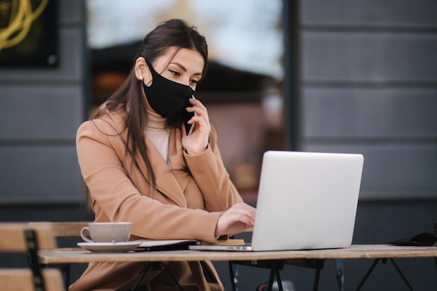 Mujer joven con mascarilla negra sentada en la terraza de un café durante la cuarentena Mujer de negocios trabajando al aire libre en cuarentena Mujer habla por teléfono Covid19