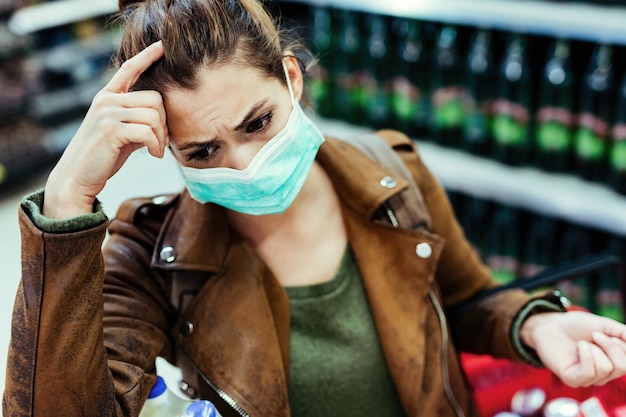 Mujer joven con mascarilla pensando y sintiéndose preocupada mientras compra en la tienda durante la pandemia de virus