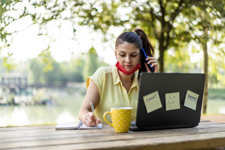 Foto mujer joven con mascarilla protectora abierta usando una computadora portátil al aire libre empresaria tomando notas mientras llama con un teléfono celular en el parque trabajo remoto nuevo concepto normal después de la pandemia de covid 19