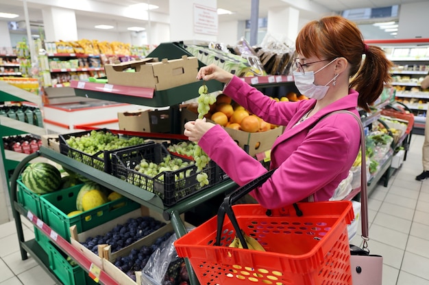 Mujer joven con mascarilla protectora, sosteniendo racimos de uvas en el supermercado