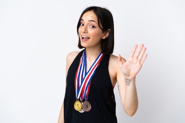 Mujer joven con medallas aislado sobre fondo blanco saludando con la mano con expresión feliz