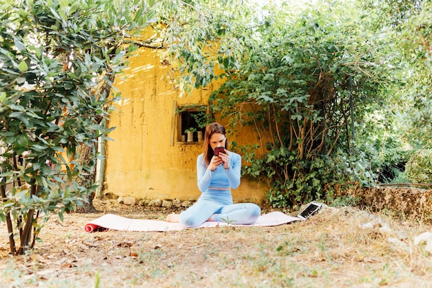 Mujer joven de mediana edad haciendo yoga en el jardín de su casa mirando el móvil. concepto de meditación, estilo de vida saludable