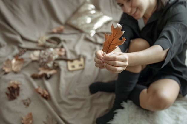 Una mujer joven con medias calientes en casa tiene una hoja de otoño en sus manos, fondo borroso.