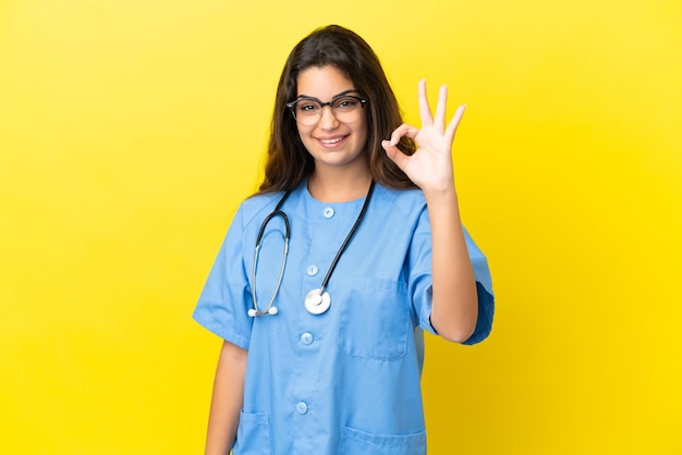 Mujer joven médico cirujano aislado sobre fondo amarillo mostrando signo ok con los dedos