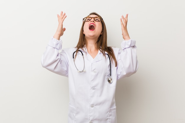 Mujer joven médico contra una pared blanca gritando el cielo, mirando hacia arriba, frustrado.