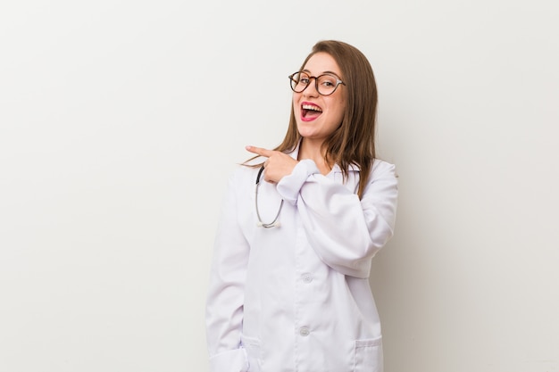 Mujer joven médico contra una pared blanca sonriendo y apuntando a un lado, mostrando algo en el espacio en blanco.