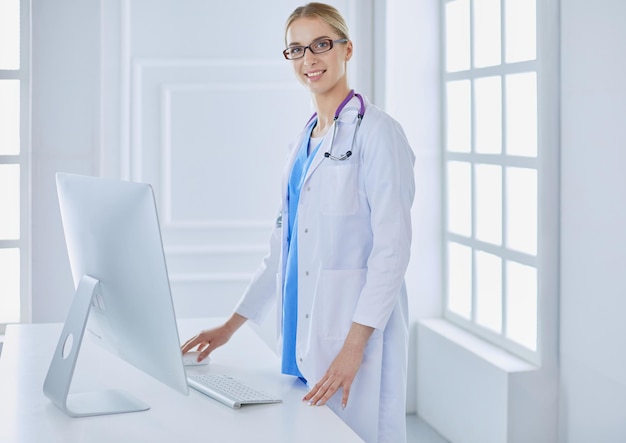 Mujer joven médico de pie junto a la mesa, aislado sobre fondo blanco.