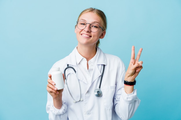 Mujer joven médico ruso que sostiene la botella de píldoras en azul que muestra el número dos con los dedos.