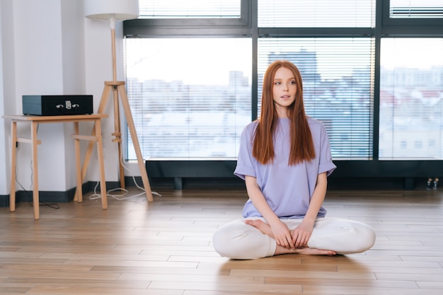 mujer joven meditando sentada en el suelo en posición de loto en el fondo de la ventana en la habitación de la oficina.