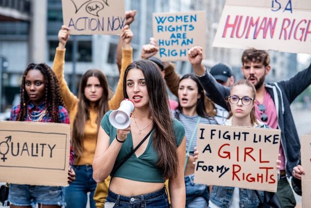 Mujer joven con megáfono liderando un grupo multirracial de personas en una huelga por la igualdad y las actividades de derechos de las mujeres en protesta en la ciudad