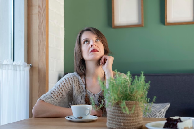 Mujer joven melancólica se sienta a la mesa en el café y mira hacia arriba Mujer de mediana edad bebe café o té en la cafetería