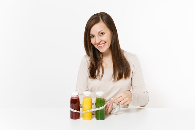 Mujer joven en la mesa con batidos de desintoxicación verdes, rojos, amarillos en botellas, cinta métrica aislada sobre fondo blanco. Nutrición adecuada, bebida vegetariana, estilo de vida saludable, concepto de dieta. Copie el espacio.