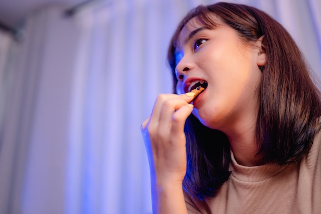 Mujer joven mientras ve la televisión, se relaja, come bocadillos y rosquillas ordenando la entrega, en casa de vacaciones. comida chatarra, comida poco saludable, riesgo de obesidad.