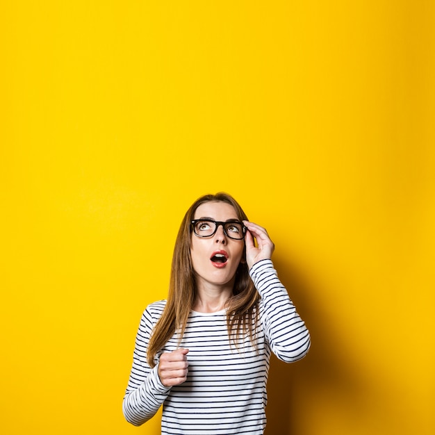 Mujer joven mira sorprendida aferrándose a las gafas sobre un fondo amarillo