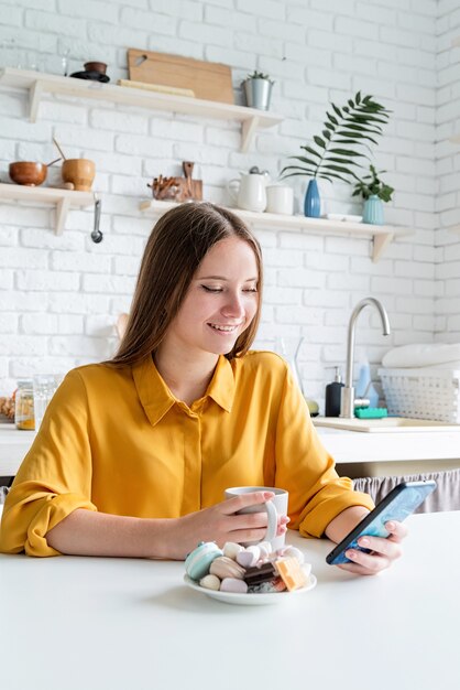 Mujer joven mira su teléfono mientras sostiene una taza sentado en la cocina