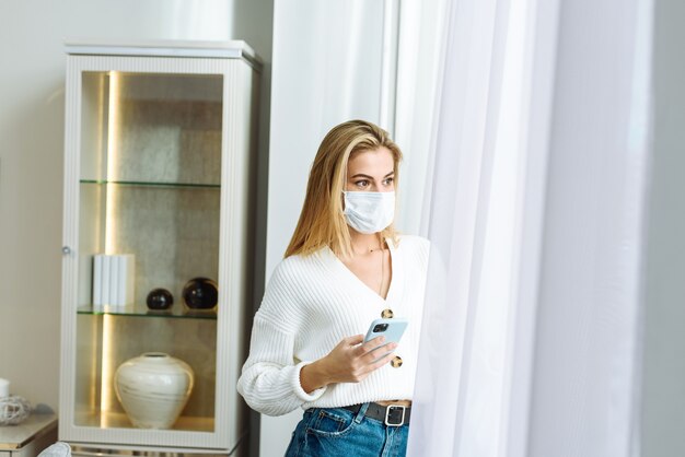 Mujer joven mira por la ventana, sostiene un teléfono móvil, cuarentena, coronavirus, búsqueda de trabajo, depresión, aislamiento.