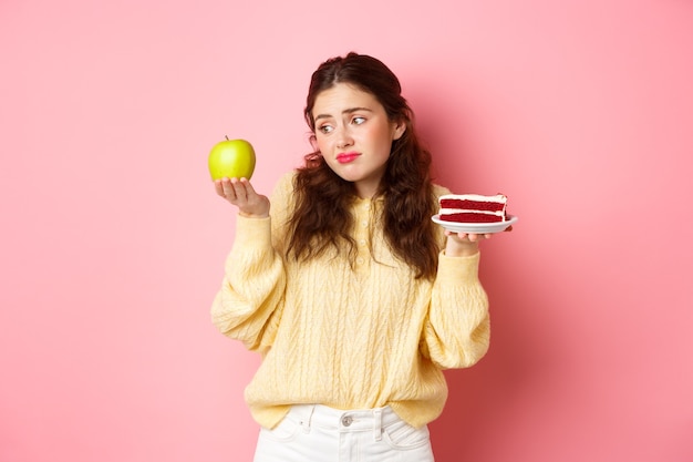 Mujer joven mirando con cara triste y decepcionada a la manzana verde mientras sostiene un trozo de delicioso pastel en otro, quiere comer postre, de pie contra la pared rosa.