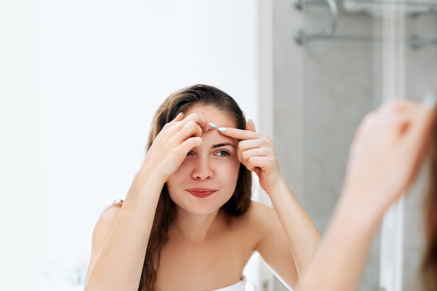 Mujer joven mirando y exprimir el acné en la cara frente al espejo. Chica fea con problemas de piel, chica adolescente con espinillas. Protección de la piel. Belleza