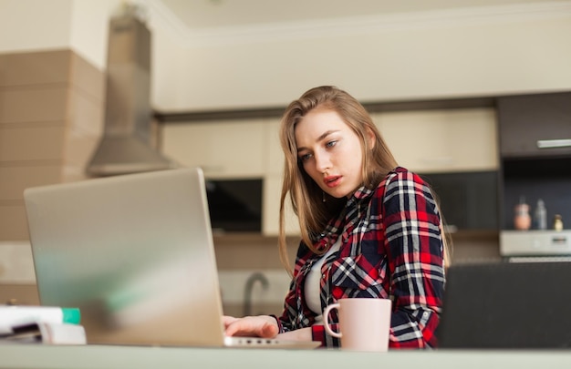 Mujer joven mirando la pantalla del portátil mientras se sienta en la mesa y bebe café en la sala de estar