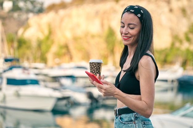 Mujer joven mirando smartphone sonriendo mientras pasea con café en la mano