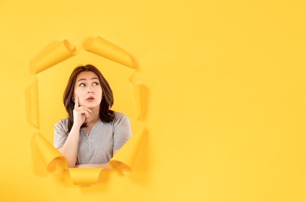 Mujer joven mirando a través de un agujero pensando en fondo de papel amarillo ventana de publicidad espía