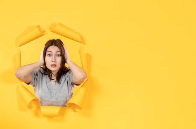 Mujer joven mirando a través de un agujero en la ventana de fondo de papel amarillo mirar