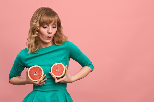 Mujer joven con mitades de pomelo en sus manos, chica alegre en verde posando en la cámara con pomelos sobre un fondo rosa, espacio de copia.