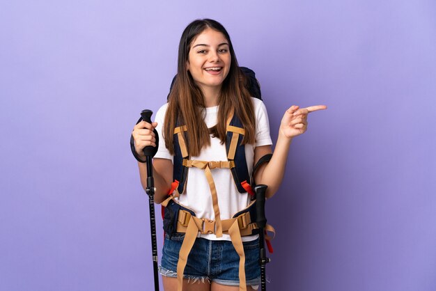 Mujer joven con mochila y bastones de trekking aislado en la pared púrpura apuntando con el dedo hacia un lado y presentando un producto