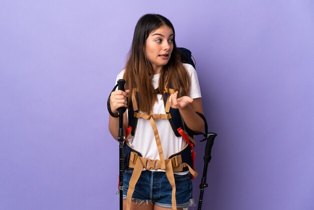 Foto mujer joven con mochila y bastones de trekking aislado sobre fondo púrpura con expresión de sorpresa mientras mira de lado