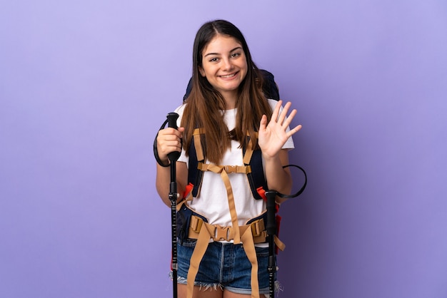 Mujer joven con mochila y bastones de trekking aislados en morado saludando con la mano con expresión feliz