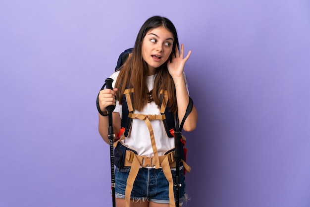 Mujer joven con mochila y bastones de trekking púrpura escuchando algo poniendo la mano en la oreja