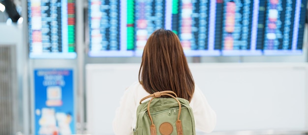 Mujer joven con mochila mirando el tablero de información de tiempo de vuelo en el aeropuerto internacional antes de registrarse en Viajes Viajes de vacaciones y concepto de transporte