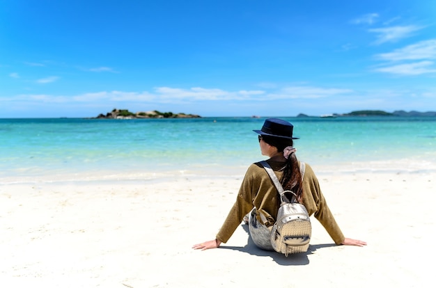 Mujer joven con la mochila que se sienta en la playa solitaria y que mira en el mar.