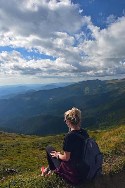 Una mujer joven con una mochila se sienta en una montaña y mira hacia el valle Vista trasera